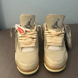 Nike Air Jordan 4 Off-White Sail - Exklusiva Nike Air Jordan 4 Off-White Sail sneakers i beige med ikoniska detaljer som genomskinliga paneler, mesh och Off-White-etiketter. Skorna har snörning, platt sula och tydlig AIR-text på sidan. Kommer med extra skosnören och klassisk Jumpman-tag.