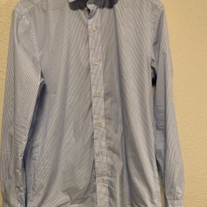 Blåvit randig skjorta från Gutteridge - Snygg blåvit randig skjorta från Gutteridge med klassisk krage och knäppning framtill. Skjortan har långa ärmar och är tillverkad i stretchigt bomullstyg för extra komfort. Perfekt för dig som gillar stilrena och fräscha plagg.