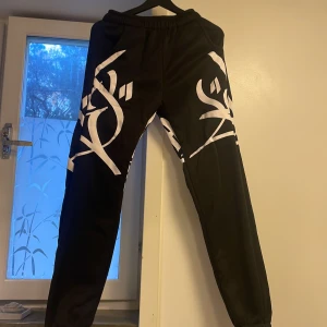 Svarta mjukisbyxor med vit print - Nya svarta mjukisbyxor med stora vita grafiska tryck längs benen. Byxorna har resår i midjan och vid bensluten för en skön och avslappnad passform. Perfekta för chill dagar eller streetwear-stil. Materialet känns mjukt och bekvämt. Oanvända. Har mindre passform än normalt, passar också dig som har stl S.