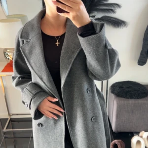 Grå lång kappa med bälte - Snygg grå kappa i ullmix med klassisk krage, dubbelknäppning och bälte i midjan. Långa ärmar med knappar vid ärmslut och slits bak för extra rörelsefrihet. Perfekt för dig som vill ha en stilren och tidlös look.Köpt för 1 år sen men bara hängt i garderoben så endast testad.   Den är helt ny. Storlek S  Köpt för 999 kr Finns inte kvar online Därför är den väldigt efterfrågad   Kan komma och bli budgivning om ingen hinner köpa den direkt❤️ 