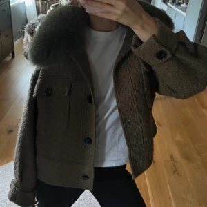 Meotine jacka - Snygg brun/grön jacka i ull med oversized grön pälsig krage. Jackan har stora svarta knappar framtill, två bröstfickor med lock och långärmad passform. Perfekt för dig som vill sticka ut med en cool och mysig look.