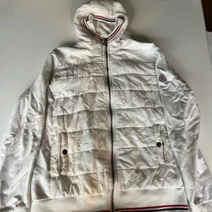 Vit cardigan från Moncler med huva - Snygg vit pufferjacka från Moncler med quiltad framsida, huva och dragkedja. Jackan har ribbade muddar och nederkant med röd, vit och blå rand. Moncler-logga på ärmen och praktiska fickor framtill. Perfekt för dig som gillar stilren och sportig look. PRIS KAN DISKUTERAS VID SNABB AFFÄR.