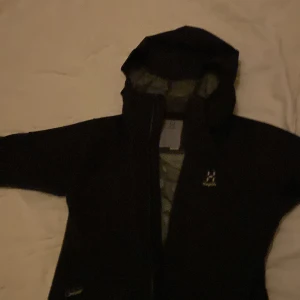 Svart vindjacka från Haglöfs med huva - Svart vindjacka från Haglöfs med hel dragkedja och justerbar huva. Jackan har meshfoder på insidan för extra ventilation och två fickor framtill. Perfekt för blåsiga dagar och outdoor-äventyr. Klassisk och stilren design med diskret logga på bröstet.