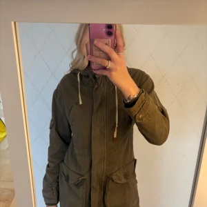 Olivgrön parkasjacka från Zara - Säljer en olivgrön parkasjacka från Zara med huva och dragkedja. Jackan har stora fickor framtill, knappar och dragkedja. Tillverkad i bomull och har en avslappnad passform. Perfekt för lager-på-lager under vår och höst. Den har även 2 dragkedjor så man kan välja hur tight den ska sitta. 