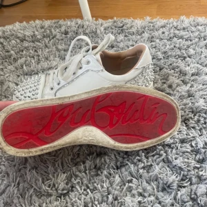 Christian Louboutin vita sneakers med nitar - Säljer ett par vita sneakers från Christian Louboutin med ikonisk röd sula och silverfärgade nitar på tån och hälen. Skorna har snörning och är tillverkade i skinn med rund tå. Perfekt för dig som vill sticka ut med en lyxig och edgy stil.
