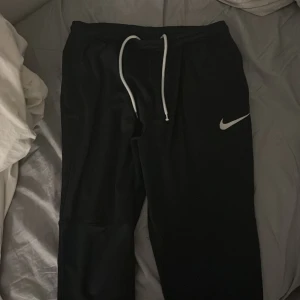 Svarta mjukisbyxor från Nike, M - Svarta mjukisbyxor från Nike med vit logga på benet och vit snörning i midjan. Byxorna har en normal passform och är tillverkade i mjukt syntetmaterial, perfekta för chill eller träning. Elastisk midja och stilren design. (Använt några få gånger) finns även en liten skada som går att ta privat, köpte dessa för 900kr med tanker på skadan på byxorna så är det lägsta 300kr.