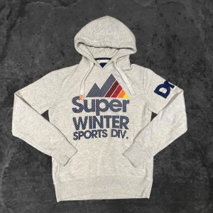Grå Superdry hoddie - Ljusgrå hoodie med tryck framtill i blått, rött, orange och gult. Tröjan har huva med snörning, ribbade muddar och ficka framtill. Snyggt grafiskt motiv med berg och texten 'Super Winter Sports Div.' samt blått tryck på ena ärmen.