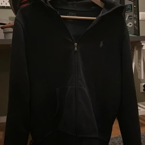  Mörk blå hoodie från Polo Ralph Lauren - Mörk blå Hoodie från Polo Ralph Lauren med dragkedja framtill och klassisk huva. Tröjan har en broderad logga på bröstet och en stor känguruficka. Perfekt för en chill och stilren look.