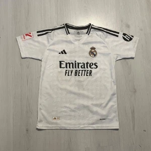 Real Madrid hemmatröja Adidas 176 - Säljer en vit Real Madrid hemmatröja från Adidas med svarta detaljer och klubbmärke på bröstet. Tröjan har korta ärmar, klassisk V-ringning och är tillverkad i lätt och ventilerande AEROREADY-material. Perfekt för dig som älskar fotboll och vill bära ditt lag.