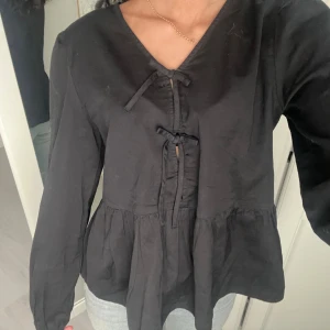 Svart blus med snörning och volang - Säljer en svart blus med v-ringning och snygg snörning framtill. Blusen har långa ärmar och en volang nedtill som ger en chill och feminin vibe. Materialet känns mjukt och lätt, perfekt att styla med jeans eller kjol. Passformen är normal och blusen är helt enfärgad.
