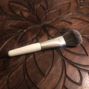 Sminkborste från CAIA F18 - Säljer en CAIA F18 sminkborste med vitt handtag och silverfärgat metallfäste. Borsten har täta, mjuka strån i svart och grått, är fluffig och är perfekt för applicering av puder, bronzer eller rouge. Snygg och stilren design som är enkel att använda.