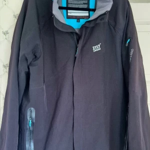 Svart vindjacka från 2117 of Sweden - Stl L - Svart vindjacka från 2117 of Sweden med diskret logga på bröstet och turkosa detaljer vid dragkedjorna i stl L.  Jackan är tillverkad i 90% polyester och 10% elastan, har hög krage och flera praktiska fickor med dragkedja. Perfekt för blåsiga dagar. Innerfodret på en av fickorna på ärmen är trasig. Lite smutsig på kragen. 