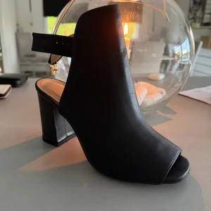 Svarta klackskor från Tata Woman - Snygga svarta klackskor från Tata Woman med öppen tå och bred klack. Skorna har en stilren design med justerbart spänne runt ankeln och är tillverkade i slätt syntetmaterial. Perfekta för dig som vill ha en modern och trendig look.