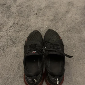 Nike Air Max 270 svarta sneakers - Svarta Nike Air Max 270 sneakers med röda detaljer och stor synlig luftbubbla i hälen. Ovandelen är i mesh för bra andning och har snörning framtill. Snygg och sportig design med röda Swoosh-loggor och 270-tryck på sidan.