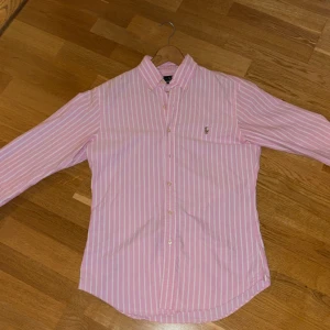 Rosa randig skjorta från Ralph Lauren - Snygg rosa skjorta från Ralph Lauren med vita vertikala ränder och klassisk broderad logga på bröstet. Slim fit-modell med lång ärm, knappar framtill och button-down krage. Perfekt för dig som gillar stilrena och fräscha plagg med lite preppy vibe.