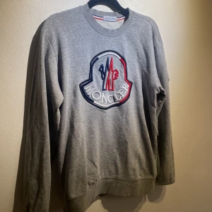 Grå Moncler sweatshirt med broderad logo - Snygg grå sweatshirt från Moncler med stor broderad logga i vitt, blått och rött på bröstet samt mindre logga på ärmen. Klassisk rund halsringning och långa ärmar. Perfekt för dig som gillar streetwear och vill sticka ut med en exklusiv vibe.