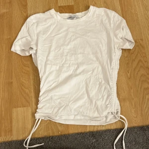 Vit snörad topp från Zara - Säljer en vit topp från Zara i storlek S. T-shirten har korta ärmar och cool snörning på båda sidorna som går att justera för en snygg passform. Materialet är mjuk bomull och modellen är normal i passningen. Perfekt för dig som gillar enkel men trendig stil.