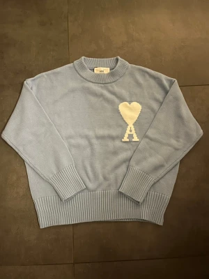 Ljusblå stickad tröja från Ami - Snygg ljusblå stickad tröja från Ami med ribbade muddar och rund hals. På bröstet finns ett stort vitt hjärta med bokstaven A under, vilket ger en unik och trendig look. Perfekt för dig som gillar stilrena detaljer och mjuka material.