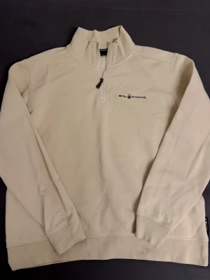 Beige Sail Racing half zip tröja - Snygg beige långärmad tröja från Sail Racing med half zip och hög krage. Diskret logga på bröstet och en liten detalj i nacken. Perfekt för dig som gillar stilren design och vill ha något bekvämt och enkelt att matcha. Materialet känns mjukt och skönt mot huden. Quarterzip, half zip, quart zip