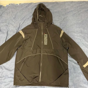 Svart vindjacka från Hoodrich L - Svart vindjacka från Hoodrich med beige detaljer på ärmarna och snygga linjer över bröstet. Jackan har huva med snörning, hel dragkedja och meshfoder på insidan. Stort Hoodrich-tryck på framsidan ger en cool streetkänsla.
