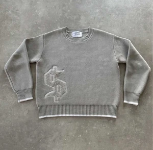 Stickad tröja! - Säljer en grå stickad tröja från Homme Studio med en stor, broderad dollarpatch på framsidan. Tröjan har rund hals, ribbade muddar och vita detaljer vid ärmslut och nederkant. Perfekt för dig som gillar streetstyle och unika detaljer.