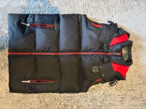 philipp plein väst  - Snygg svart pufferväst från 4P 7/8 med röda detaljer och dragkedjor. Västen har huva, två fickor med dragkedja och coola paneler i rött vid axlarna. Perfekt för lager-på-lager och streetstyle. Passar dig som gillar sportig och modern look.