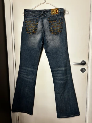 Blå bootcut jeans från Baby phat  - Blå bootcut jeans från Baby Phat med gult broderi på bakfickorna. Lågmidjade och stretchiga i tyget. Storlek rakt över midjan 36 och innerbenslängd är 83. Dm vid frågor! 