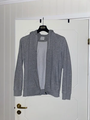 Grå hoodie med dragkedja från Jack & Jones - Snygg grå hoodie från Jack & Jones med dragkedja framtill och två fickor. Tröjan har huva med snörning och är tillverkad i mjuk bomullsmix. Perfekt för chill dagar och enkel att matcha med jeans eller joggers.