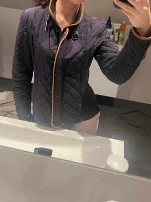 Mörkblå quiltad jacka med bruna detaljer - Snygg mörkblå quiltad jacka med bruna kantdetaljer och lång ärm. Jackan är storlek M, men skulle säga att den sitter som storlek. S för referens är jag 162 cm