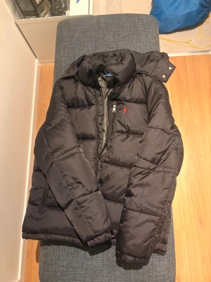 Svart pufferjacka från Polo Ralph Lauren - Svart pufferjacka från Polo Ralph Lauren med röd broderad logga på bröstet. Jackan har huva, dragkedja med Polo-detalj och två sidofickor. Materialet är syntet och jackan är quiltad för extra värme. Perfekt för kalla dagar och har en clean, stilren look.