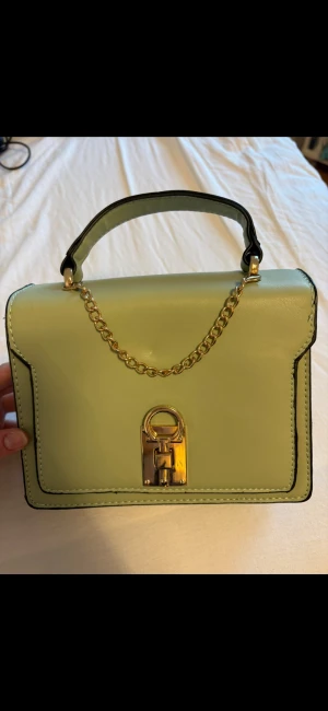 Grön handväska - Denna handväskan är grön med ”guldiga detaljer”. Det finns ett långt band med till handväskan. Varumärket är okänt. Den är aldrig använd.