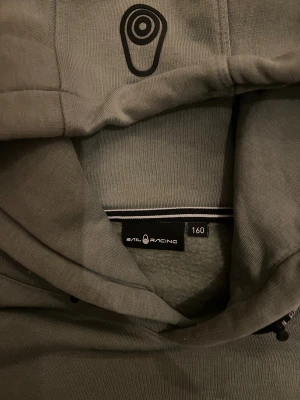 Grön hoodie från Sail Racing - Snygg grön hoodie från Sail Racing med svart logga på huvan och diskret Sail Racing-märke på magen. Tröjan har huva med snörning, stor magficka och långa ärmar. Perfekt för chill eller häng med kompisar.