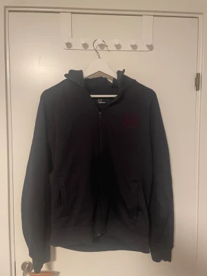 Svart bomberjacka från Fred Perry - Svart bomberjacka från Fred Perry med dragkedja framtill och två sidofickor. Jackan har hög krage och ett diskret rött Fred Perry-emblem på bröstet. Tillverkad i mjuk bomull, perfekt för dig som gillar stilren och enkel design.