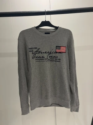 Grå sweatshirt från J.F.K med tryck - Snygg grå sweatshirt från J.F.K med broderad amerikansk flagga och texten 'American Ocean Crew' på bröstet. Tröjan har rund hals, långa ärmar och är gjord i mjuk bomull. Den är i fint skick!