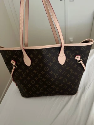 Louis Vuitton Neverfull monogramväska - Säljer en klassisk Louis Vuitton Neverfull med ikoniskt monogrammönster i brunt och beige. Väskan har ljusa skinnhandtag, guldiga metalldetaljer och randigt innerfoder. Perfekt storlek för att få plats med allt du behöver och snygg att bära på axeln.