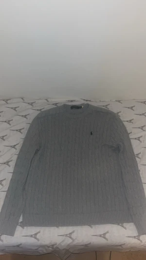 Grå stickad tröja Polo Ralph Lauren XL - Snygg grå stickad tröja från Polo Ralph Lauren i storlek XL. Tröjan har klassiskt kabelstickat mönster, rund hals och lång ärm. Diskret broderad logga på bröstet. Perfekt för dig som gillar stilrena och tidlösa plagg.
