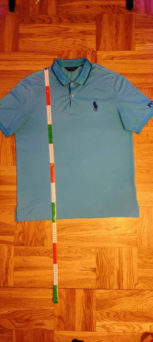 Blå pikétröja från Polo Ralph Lauren - Snygg blå pikétröja från Polo Ralph Lauren med klassisk krage och två knappar. Tröjan har broderad logga på bröstet och detaljer på båda ärmarna, inklusive texten 'POLO' och en trofé. Materialet är mjuk bomull som andas och passar perfekt till en chill stil.