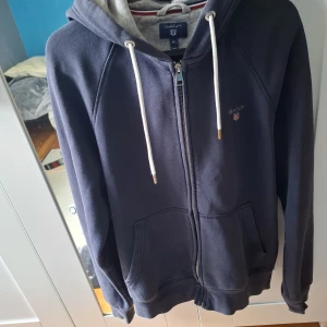 Mörkblå hoodie med dragkedja från GANT - Säljer en mörkblå hoodie från GANT med dragkedja och vit snörning i huvan. Tröjan har två fickor framtill och en liten broderad GANT-logga på bröstet. Insidan av huvan är ljusgrå och materialet är mjukt och bekvämt.