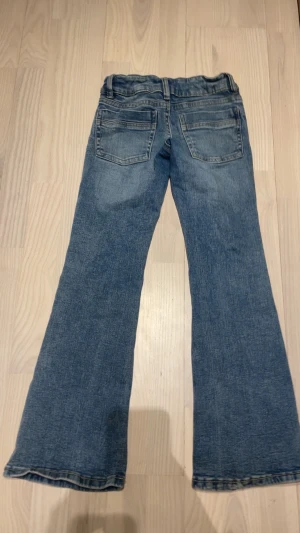 Blå bootcut jeans från Gina Tricot - Ett par klassiska blå jeans från Gina Tricot i storlek 146. Jeansen har bootcut-modell med utsvängda ben och femficksdesign. Tillverkade i jeansmaterial med snygga slitningar och detaljerade sömmar. Perfekta för en avslappnad och trendig look.