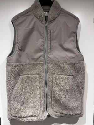 Beige väst med dragkedja - Säljer en beige teddyväst från studio total  med mjuk fleece och två stora fickor framtill. Västen har hög krage och dragkedja hela vägen. Perfekt att slänga över hoodien för extra värme och chill stil. 