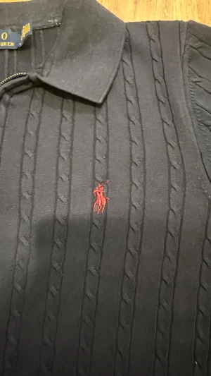 Marinblå pikétröja Polo Ralph Lauren - Snygg marinblå pikétröja från Polo Ralph Lauren med kort ärm och half zip-dragkedja. Tröjan har kabelstickat mönster och röd broderad logga på bröstet. Klassisk krage och ribbade muddar ger en clean och stilren look. Perfekt för dig som gillar preppy stil.