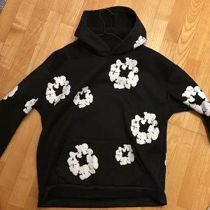  Denim Tears hoodie  - Svart hoodie från Denim Tears med stora vita blommor tryckta över hela tröjan, inklusive huvan. Tröjan har klassisk känguruficka framtill och en skön huva. Perfekt för dig som gillar streetwear med unik design.