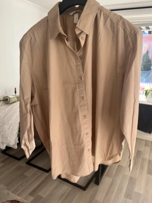 Beige skjorta från H&M i bomull - Snygg beige skjorta från H&M i storlek M. Skjortan har klassisk krage, knappar hela vägen och långa ärmar med knapp vid manschetten. Tillverkad i mjuk bomull och har en loose passform som ger en chill vibe.