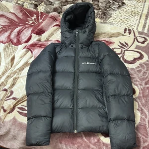 Svart dunjacka från Sail Racing - Svart dunjacka från Sail Racing med huva och diskret logga på bröstet. Jackan har puffad design, dragkedja framtill och innerficka med zip. Perfekt för kalla dagar och har en stilren look som funkar till det mesta.