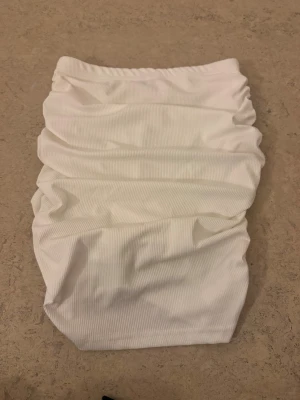 Vit ribbad kortkjol från SHEIN XS - Supersnygg vit kortkjol från SHEIN i storlek XS. Kjolen har ribbad struktur och är gjord i stretchigt syntetmaterial. Den har rynkade detaljer längs sidorna som ger en cool och trendig look. Perfekt att matcha med crop tops eller hoodies. Har även en inbyggd kjol