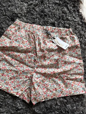 Blommiga pyjamasshorts från Change XS - Säljer ett par blommiga pyjamasshorts från Change i storlek XS. Shortsen har en ljus bottenfärg med rosa, blå och röda blommor, elastisk midja och är gjorda i mjuk bomull. Perfekta för chill dagar hemma eller mysiga kvällar.