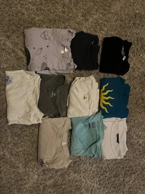 10-pack t-shirts i olika färger och tryck - Säljer ett paket med 10 t-shirts i olika färger och stilar. Här finns bland annat en ljuslila tee med space-tryck och glitterdetaljer, en blå med gul sol, svart, grå, beige och flera vita. Några har tryck med blommor eller text. Alla är klassiska kortärmade t-shirts i mjuk bomull.