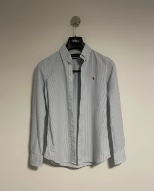 Randig skjorta från Polo Ralph Lauren - Snygg skjorta från Polo Ralph Lauren med klassiska blå och vita ränder. Skjortan har lång ärm, knappar framtill och ikoniska broderade loggan på bröstet. Tillverkad i mjuk bomull och har en normal passform med krage och manschetter.