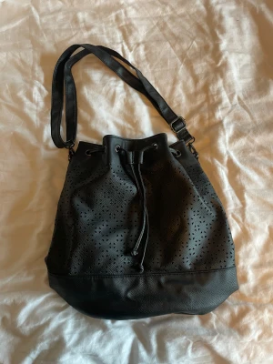 Svart axelväska - Vintage Svart medel stor väska i skinnimitation med justerbar axelrem och utskurna detaljer i triangelform. Väskan har snörning upptill för att stänga och en mjuk, rundad form som gör den både praktisk och trendig. 