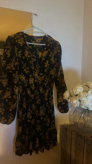 Svart blommig kortklänning från SHEIN - Supersöt svart kortklänning från SHEIN med gula blommor, v-ringning och långa puffärmar. Klänningen har smock i midjan och volangkant nertill för en extra feminin touch. Perfekt för dig som gillar romantiska vibes och snygga detaljer.
